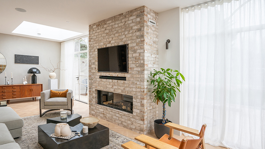 Contemporary living room featuring VENESTRE bespoke sheer curtains, allowing natural light to filter through floor-to-ceiling windows. The custom window treatments add elegance and softness, complementing the exposed brick fireplace and modern interior design.