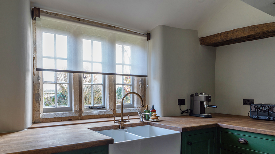 Cotswolds kitchen featuring VENESTRE made-to-measure sheer roller blind, installed above a farmhouse sink and original stone windows. The custom window treatment provides privacy and soft daylight, blending seamlessly with rustic architectural features.