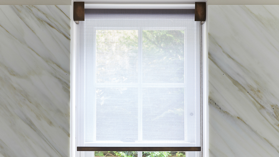 Close-up of VENESTRE roller blind in sheer fabric with antique bronze hardware, installed inside a recess of a window. 