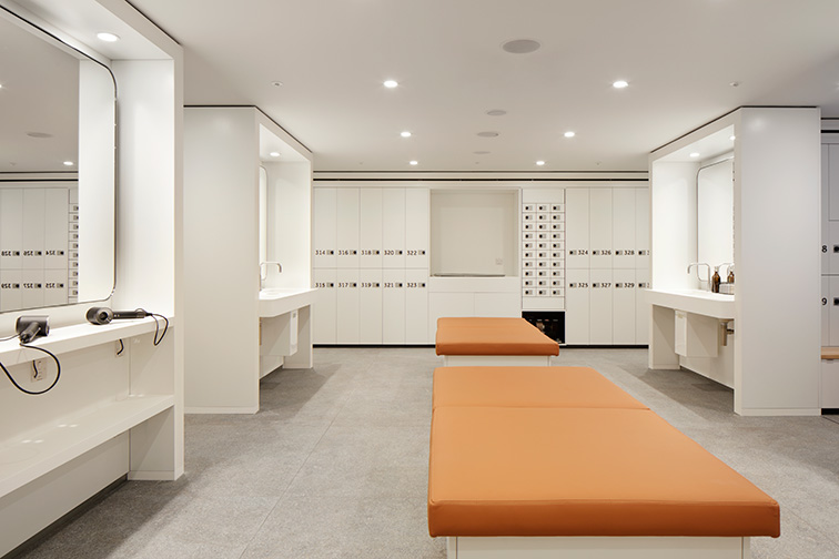 Vanity stations to the left of the image, a bench is located in the middle and lockers are seen at the back of the room