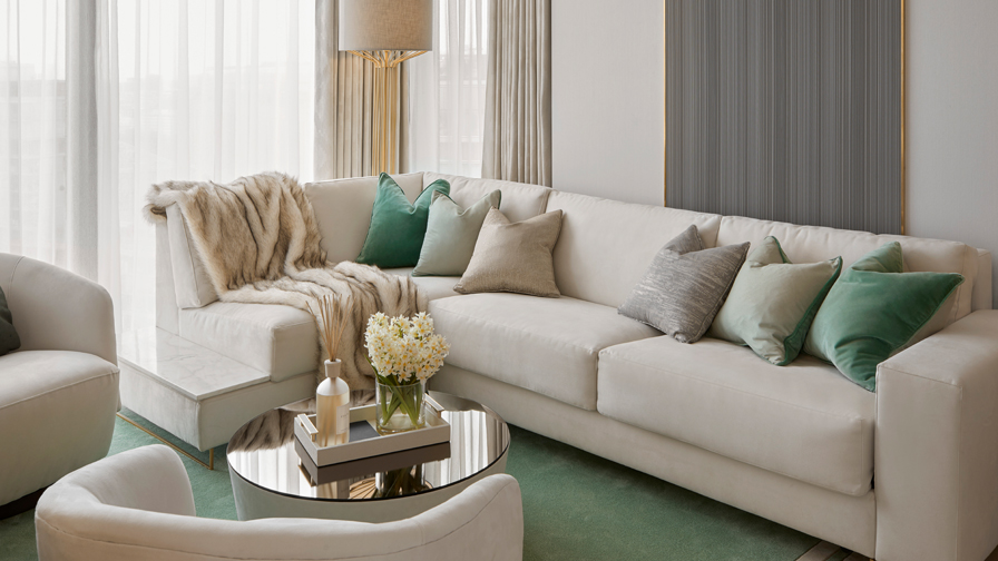 Luxury modern living room with ivory sectional sofa, soft green and neutral accent cushions, marble-topped side panels, fur throw, and mirrored coffee table with fresh flowers