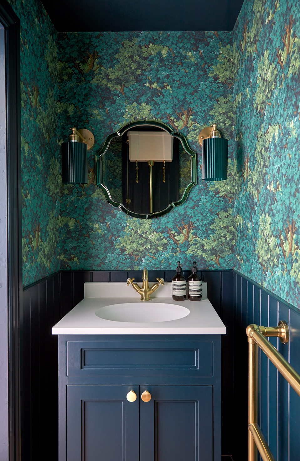 A small cloakroom with rich teal and navy tones. The walls feature a leafy patterned wallpaper with dark panelling below, complemented by brass fixtures and lighting. A scalloped mirror and navy vanity with a white countertop complete the bold, elegant look.