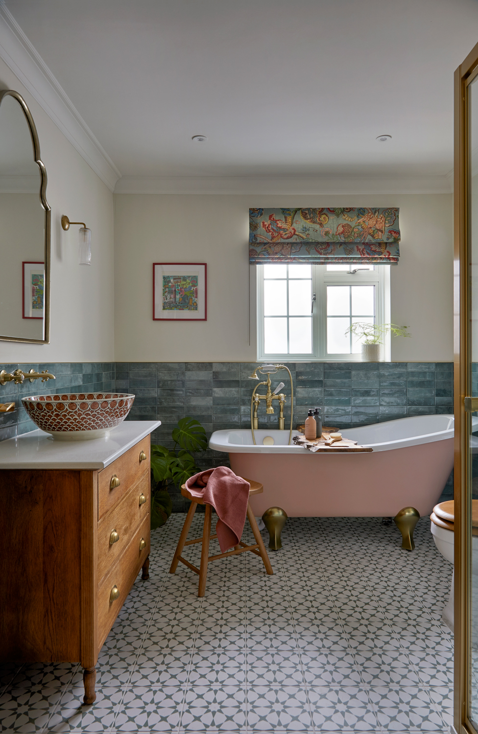 A stylish bathroom with a soft pink freestanding bath, brass fittings, and patterned floor tiles. Green glazed wall tiles and a colourful Roman blind add character, while a wooden vanity with a patterned basin brings warmth to the space.