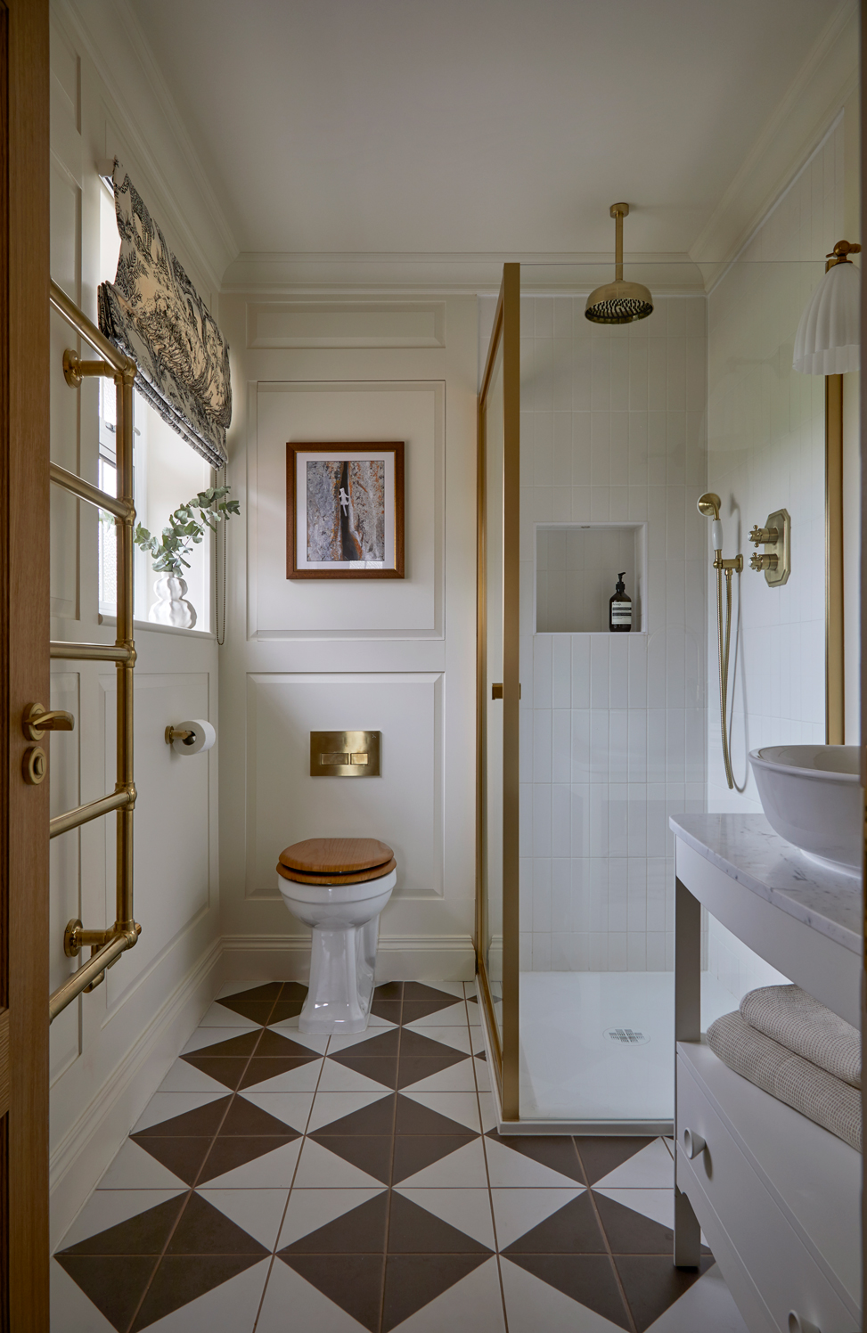 A refined shower room with cream panelling, brass fittings, and geometric floor tiles in brown and white. It features a glass shower enclosure, a wooden toilet seat, and a marble-topped vanity, with a patterned Roman blind adding a touch of elegance.