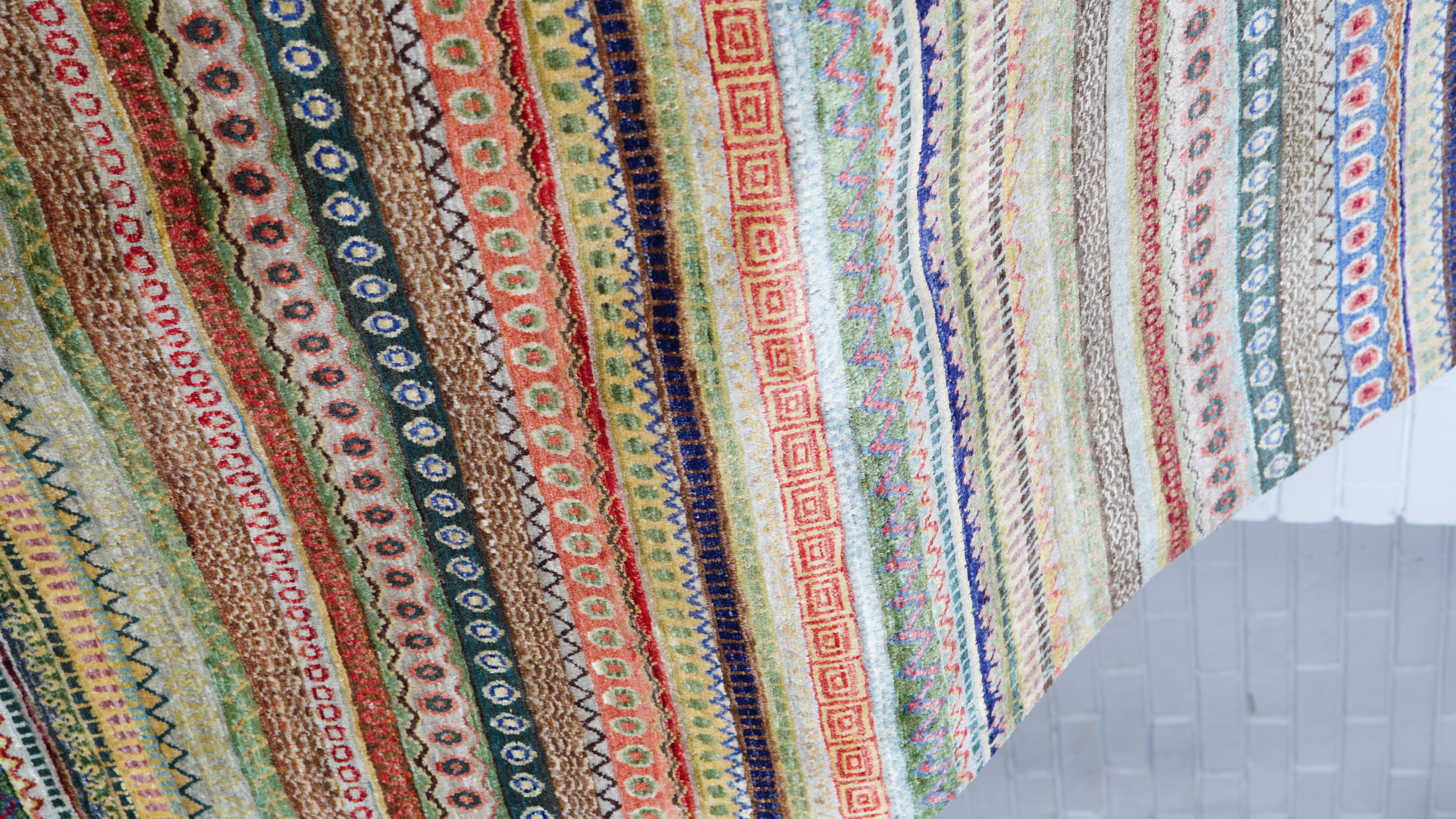 A close-up, angled view of a hand-knotted Afghan Khurjeen rug, featuring multiple horizontal stripes of various intricate geometric patterns in a multicoloured, earthy palette.