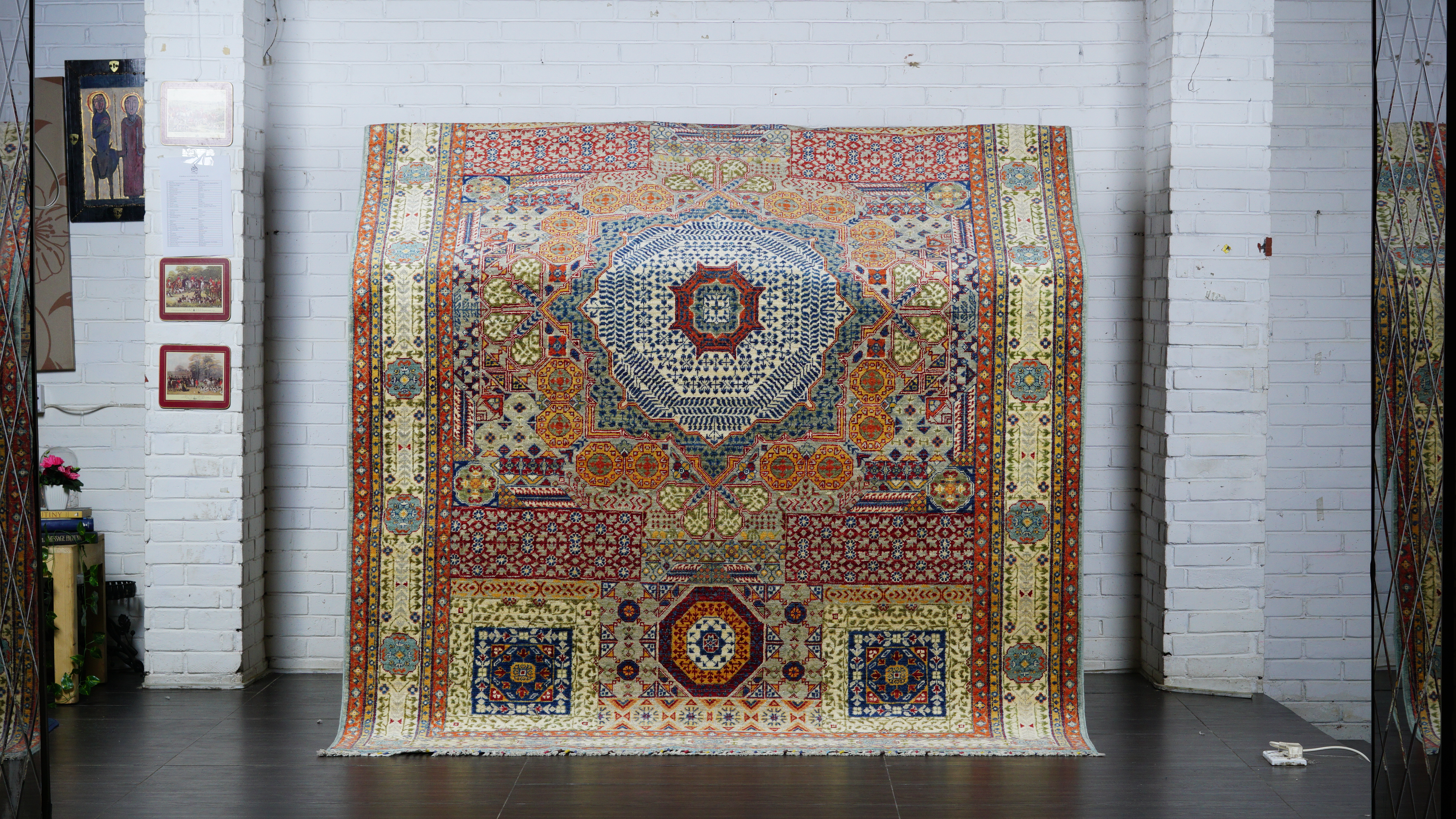 A large, hand-knotted Afghan Mamluk rug displayed vertically against a white brick wall, featuring a highly intricate geometric pattern with a central medallion and multiple borders in red, blue, and gold tones.