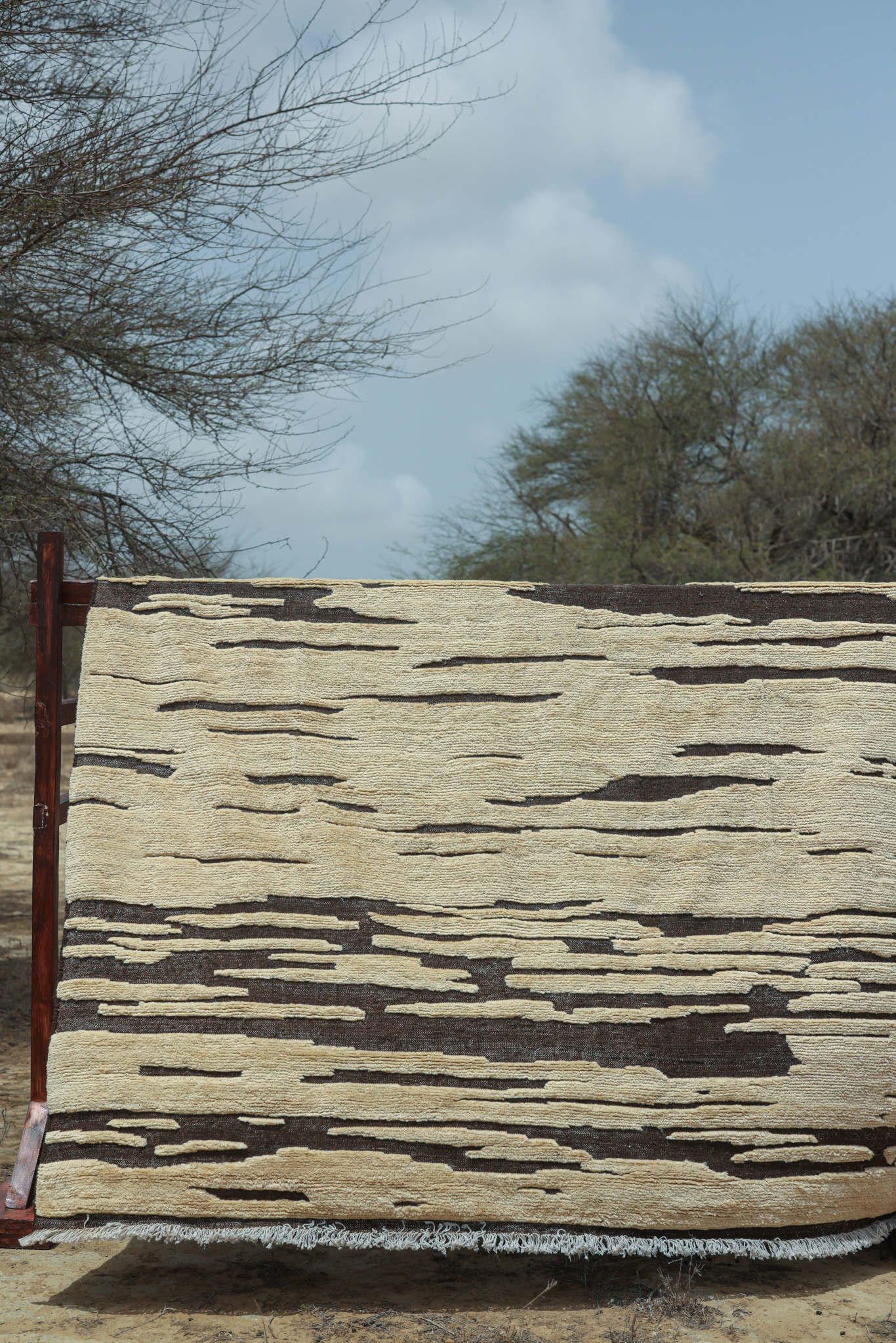 A hand-knotted rug from the Imaco Originals collection, featuring a high-low pile with a "broken stripe" abstract pattern in natural undyed cream and dark brown wool, hung outdoors for display.
