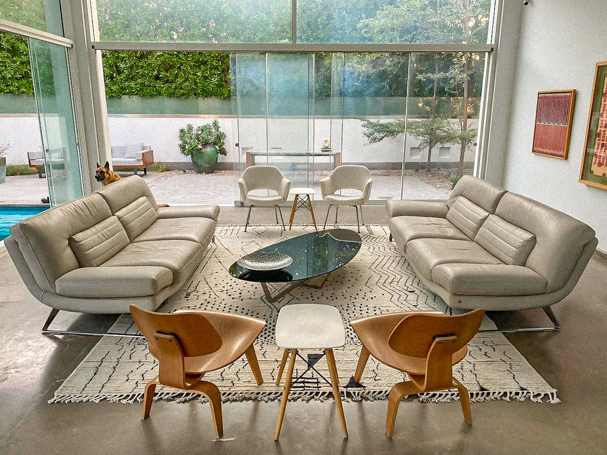 A spacious, modern living room with floor-to-ceiling windows, two grey leather sofas, and iconic wooden chairs, all arranged on a large, hand-knotted, cream-coloured Moroccan rug with a black geometric pattern.