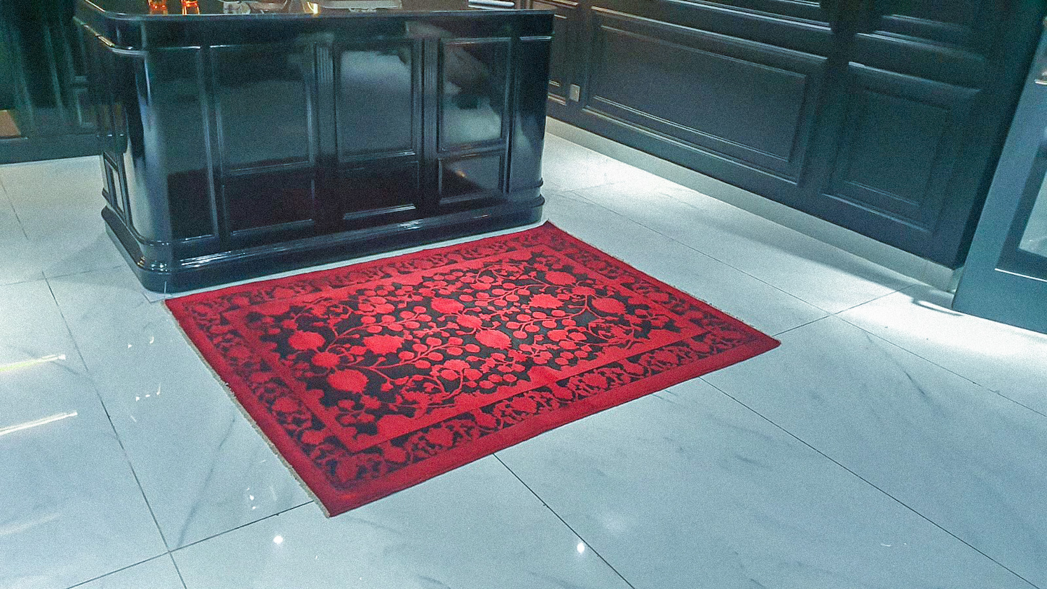 A hand-knotted, red-and-black rug with a floral pattern, placed on a white marble floor in front of a glossy black panelled counter.