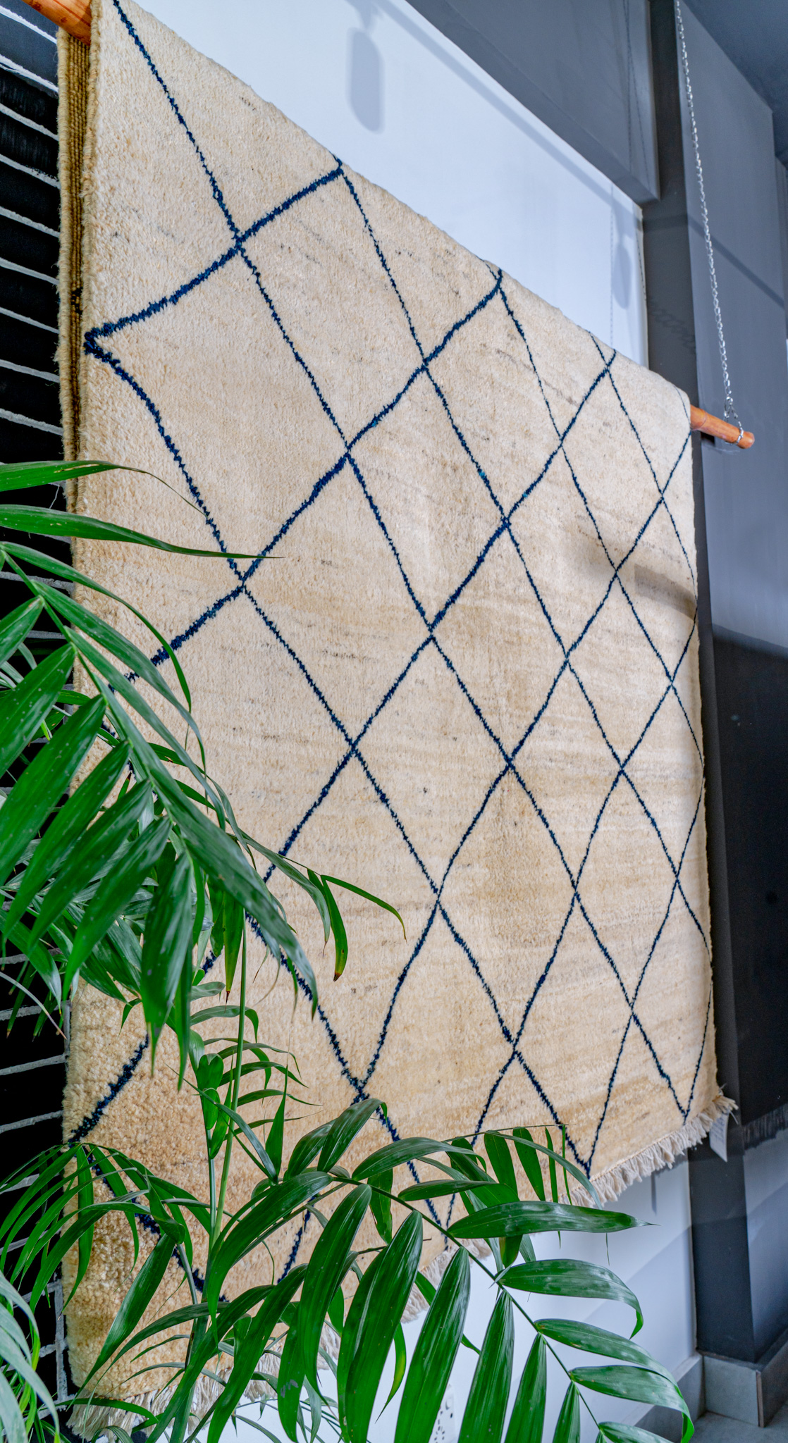 A plush, hand-knotted, cream-coloured Moroccan-style rug with a dark blue diamond lattice pattern, hung for display with a green plant in the foreground.