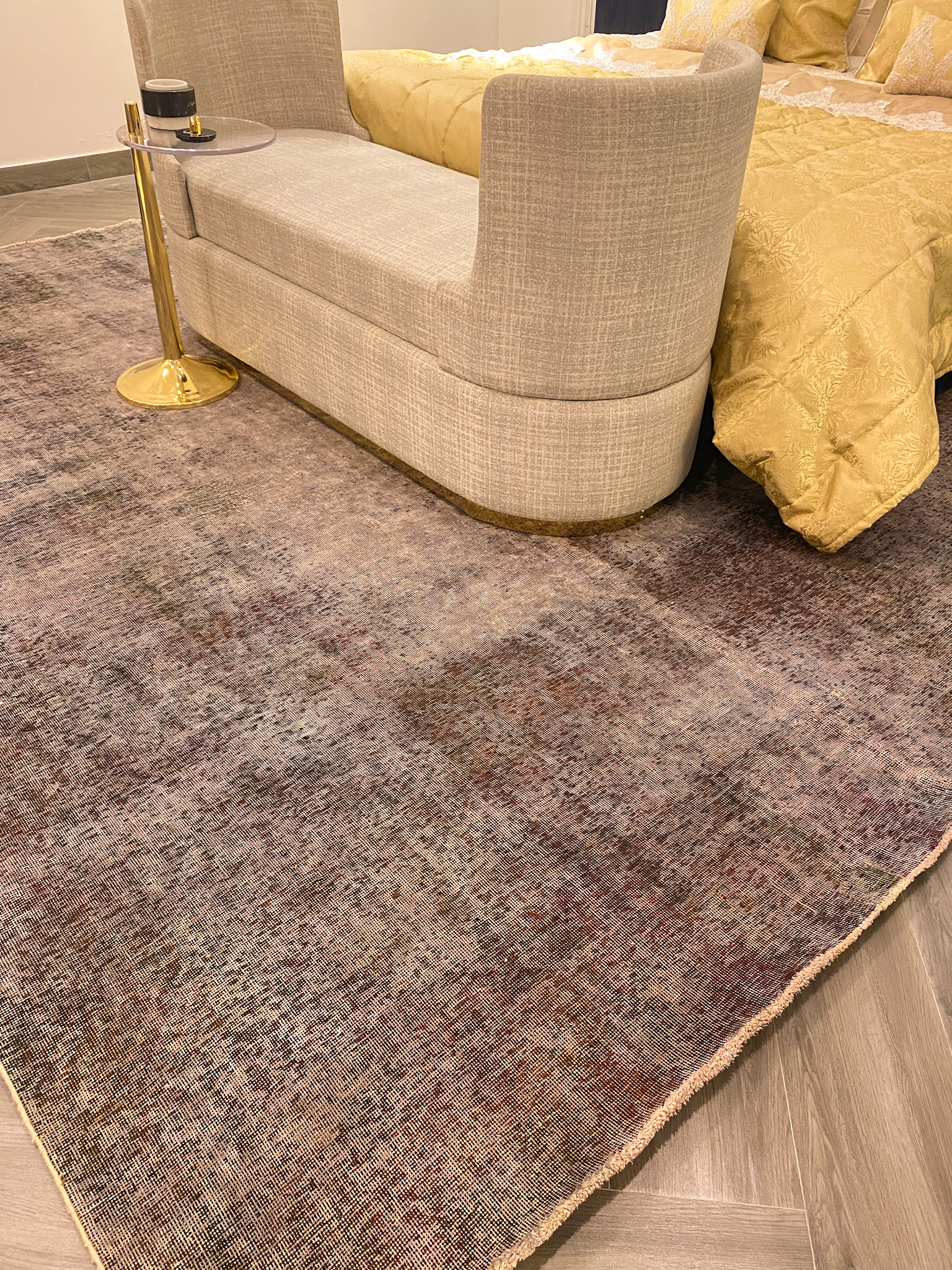 A close-up of a large, overdyed Persian rug with a heavily distressed, vintage look, in faded, purplish-brown tones, placed on a light wood floor at the foot of a bed.