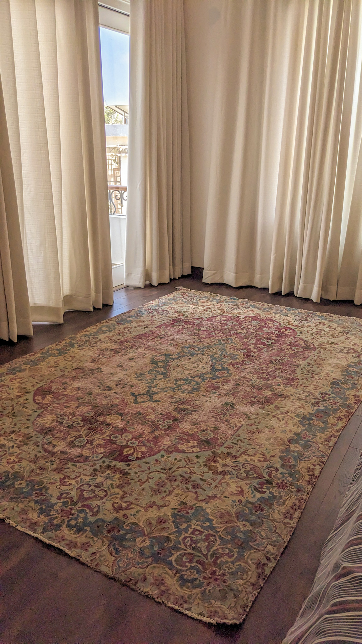 A large, overdyed Persian rug in a bedroom, placed on a dark wood floor. The rug has a distressed, vintage appearance with a faded floral pattern in rose, blue, and cream tones.