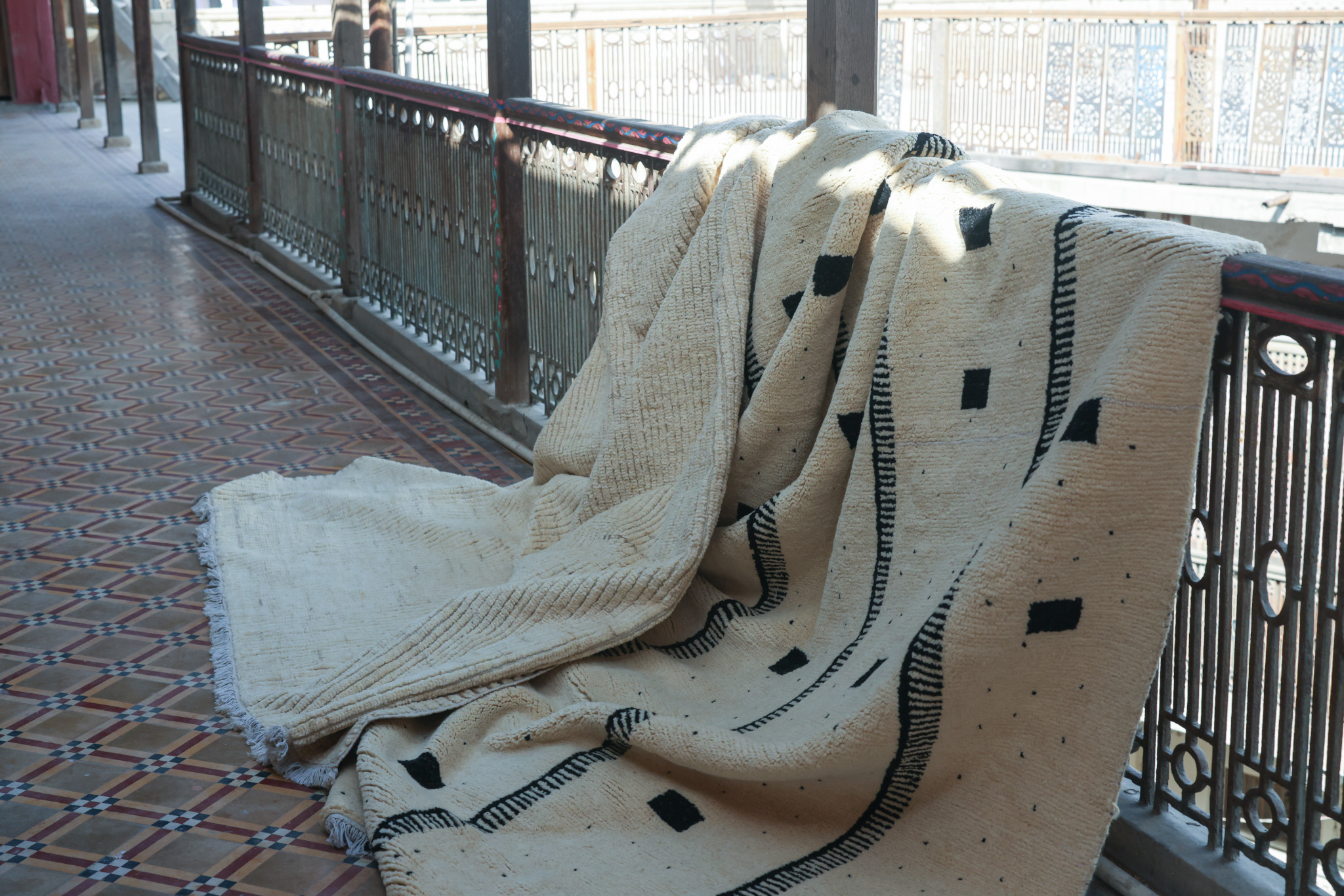 A hand-knotted, high-pile rug from the Imaco Originals collection in natural undyed cream wool, featuring a simple, bold pattern of black squares and curved lines, draped over a railing on a tiled balcony.