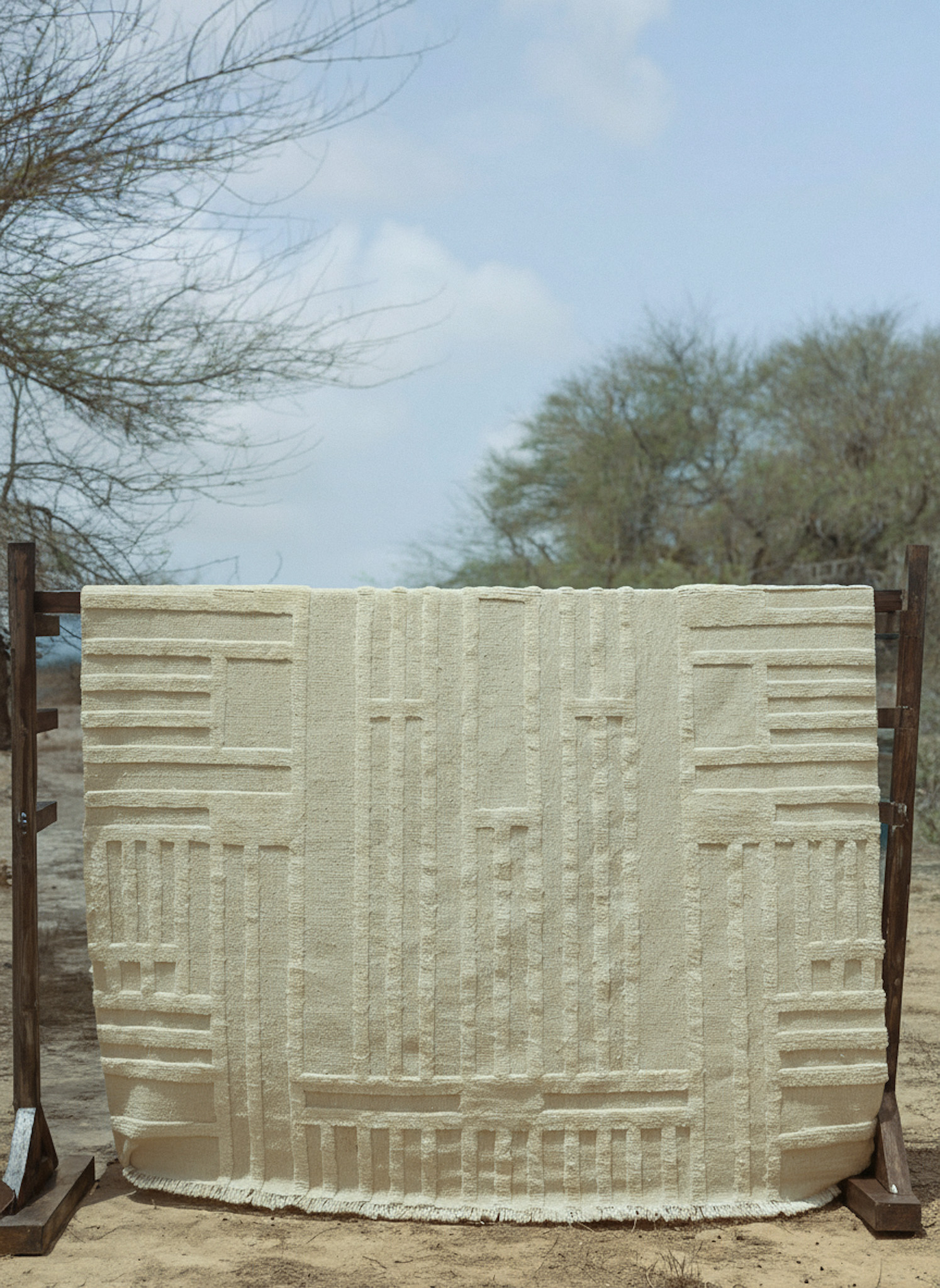 A hand-knotted rug from the Imaco Originals collection, named 'Veluxia', featuring a high-low "sculpted" pile in an abstract, geometric block pattern, all in a single, natural, undyed cream wool, hung outdoors for display.