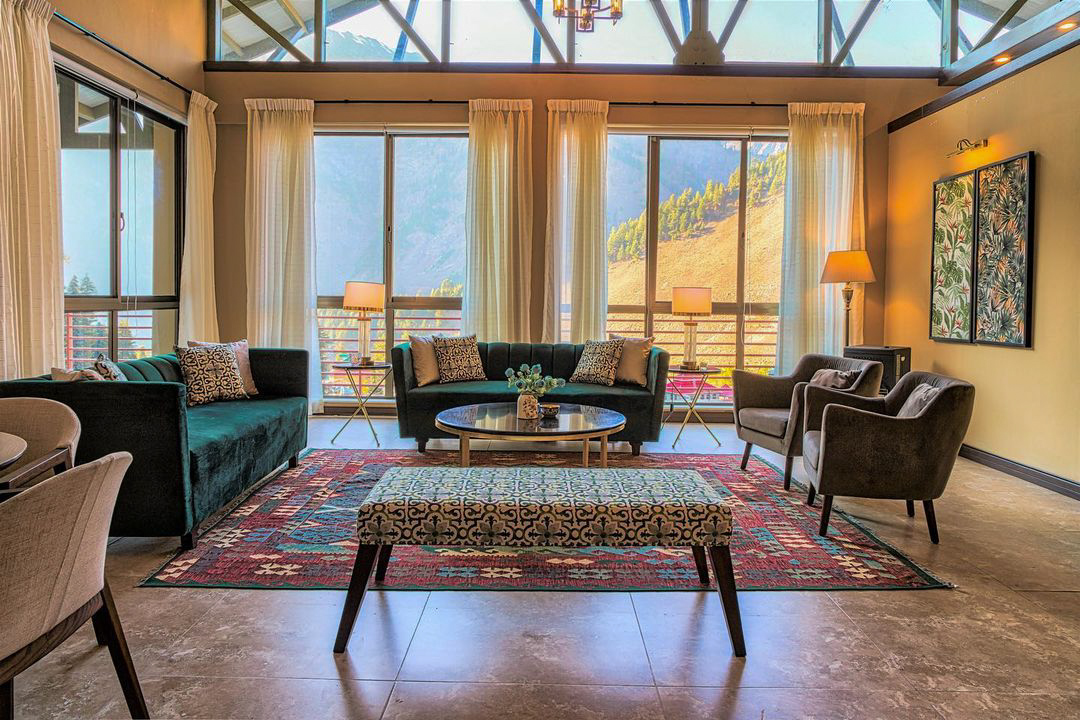 A sunlit mountain retreat lounge featuring a large, geometric kilim rug in red and blue, dark green sofas, and large windows with a view of a sunny mountain landscape.