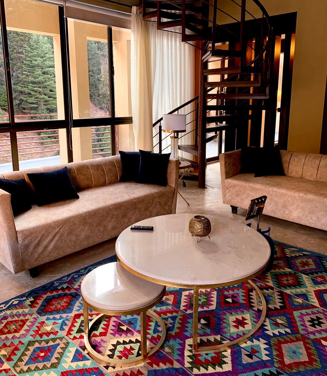 A vibrant, geometric kilim rug in blue, red, and purple, placed under modern beige sofas and a white marble nesting table, with a metal spiral staircase in the background.