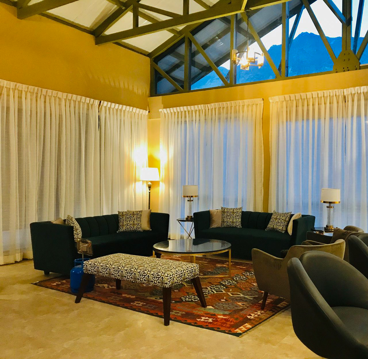 A large lounge in a mountain retreat at dusk, featuring a geometric red and orange kilim rug, two dark green sofas, and a patterned bench, all under a high, exposed-truss ceiling.