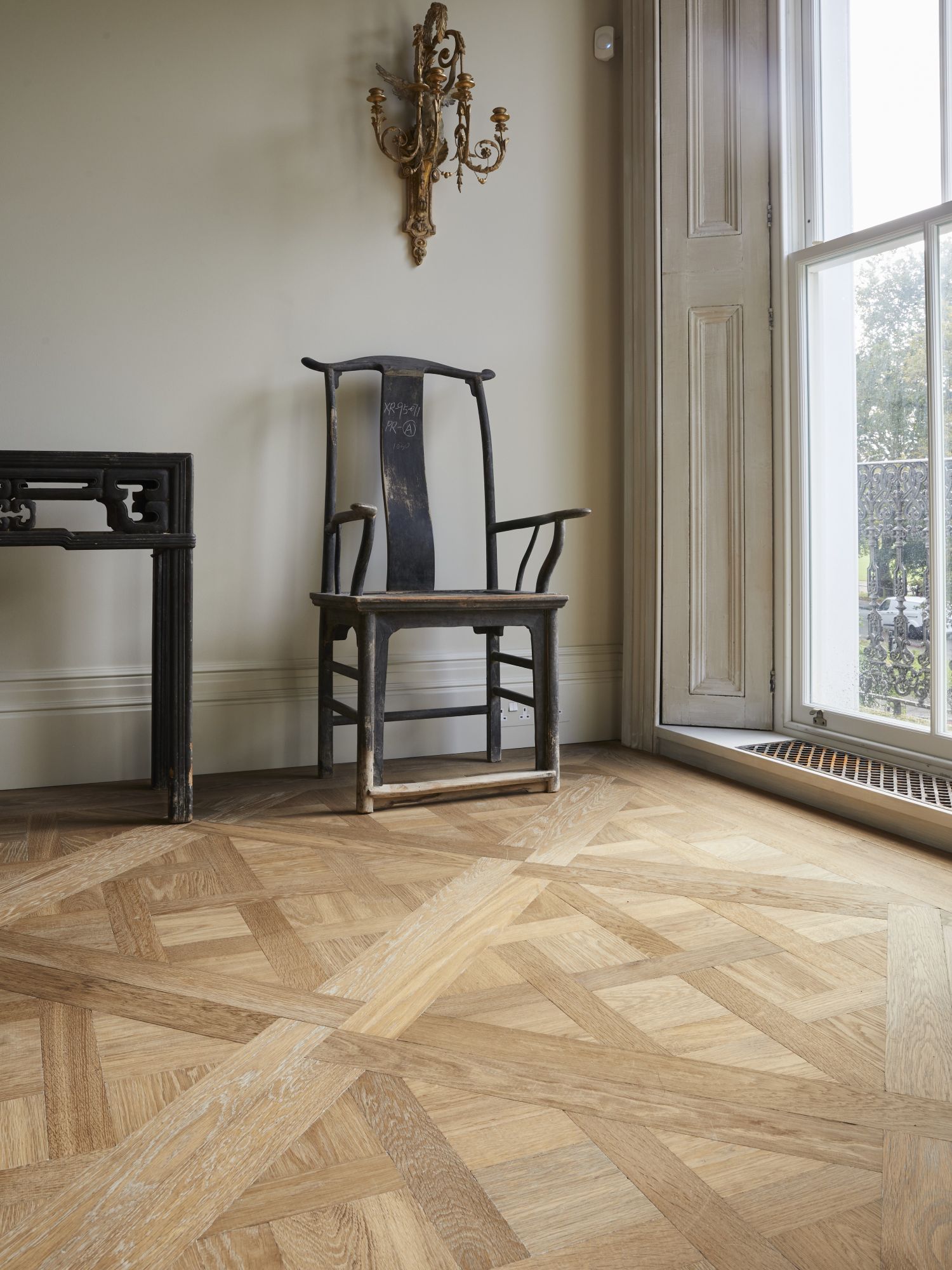 The corner of a room featuring Versailles parquet and an Asian antique chair. On the left a glimpse of a  ceiling to floor window. 