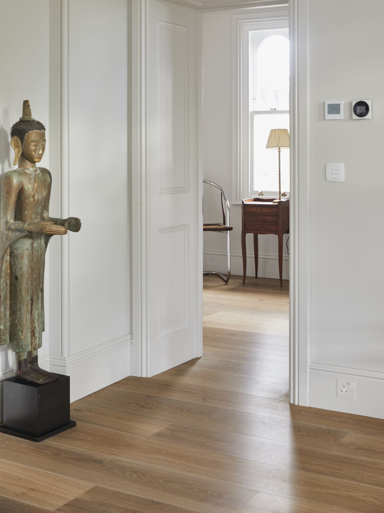 A view from one interior into the next room with white walls and a neutral coloured oak floor flowing throughout , a tall Asian statue stands on the lefthand side.