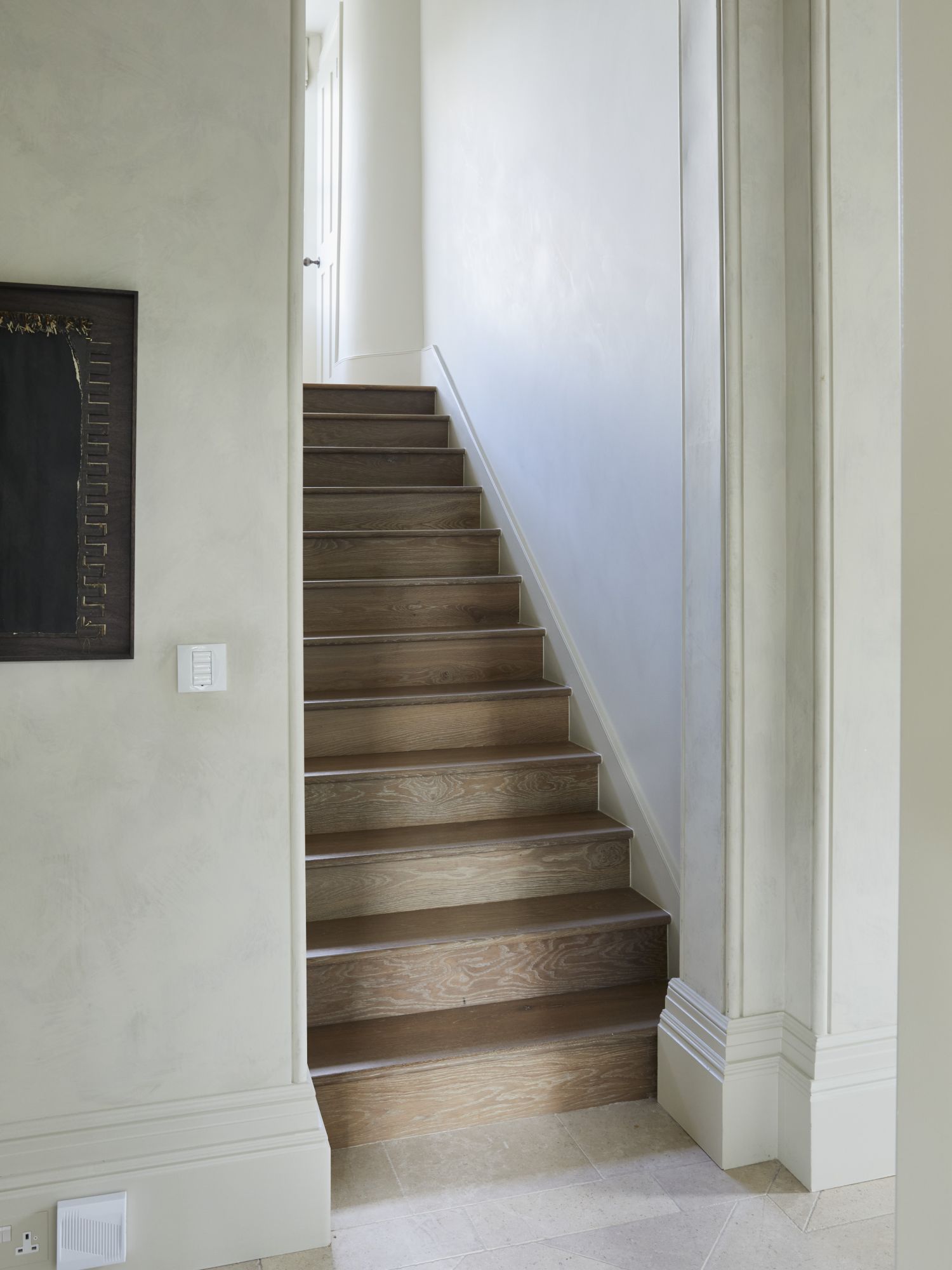 The view from a white hallway with a stone floor onto a staircase clad in oak.