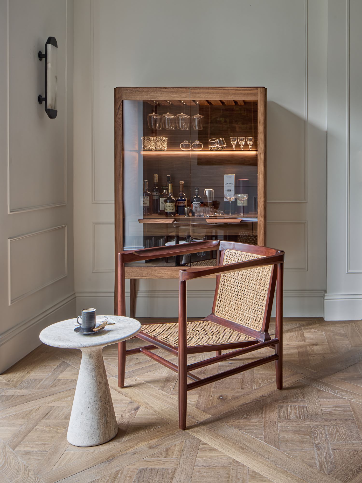 A marble round side table with cup and saucer next to a rattan mid-century chair on top of a Versailles parquet oak floor and glass side board in th back ground