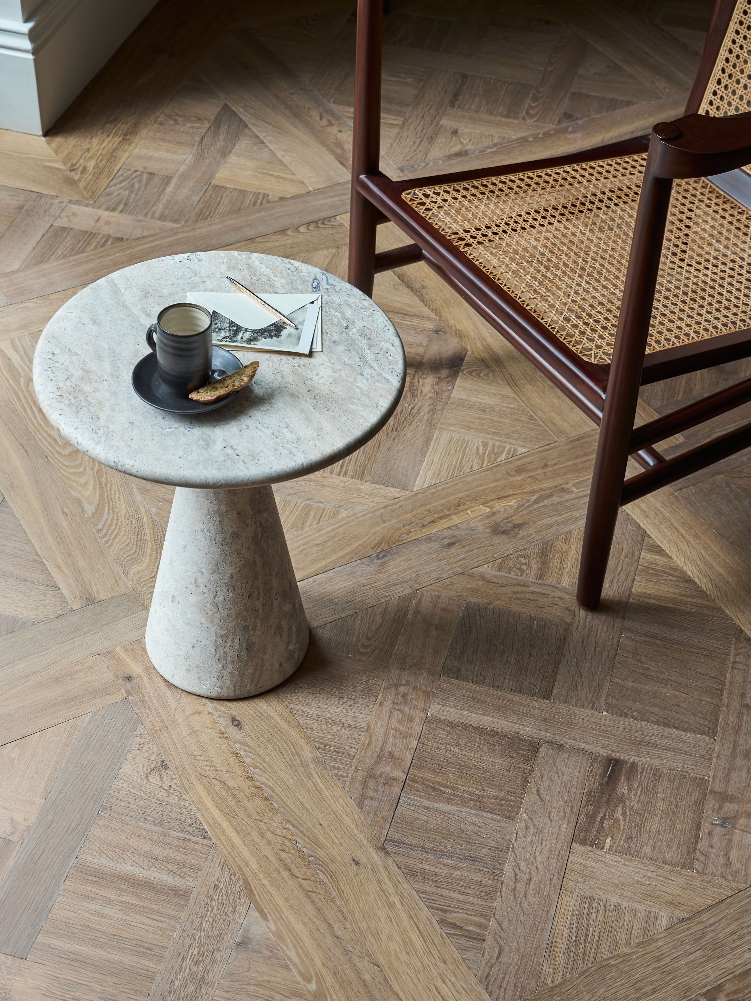 Top down view of a marble round side table with cup and saucer next to a rattan mid-century chair on top of a Versailles parquet oak floor.
