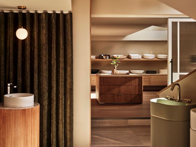 Two sinks in front corners, one with white basin, silver tap and curved wooden base, the other an olive colour with gold tap and curved base. Selection of white sinks in the background on shelves.