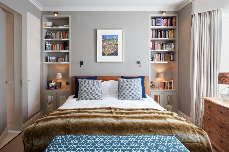 A double bed with a brown blanket and 2 grey pillows, with bookshelves either side and long cream curtains.