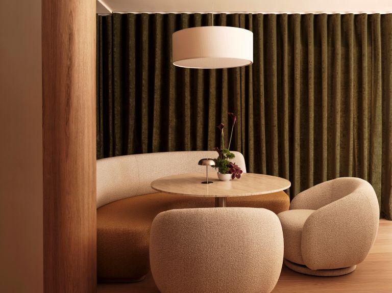 Dark curtains, with a round table in front with 2 sandy coloured curved chairs and a sofa seat.