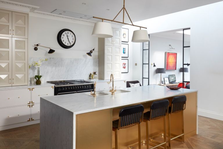 A kitchen with 3 bar stools at a central island, a clock and a sink with golden taps.